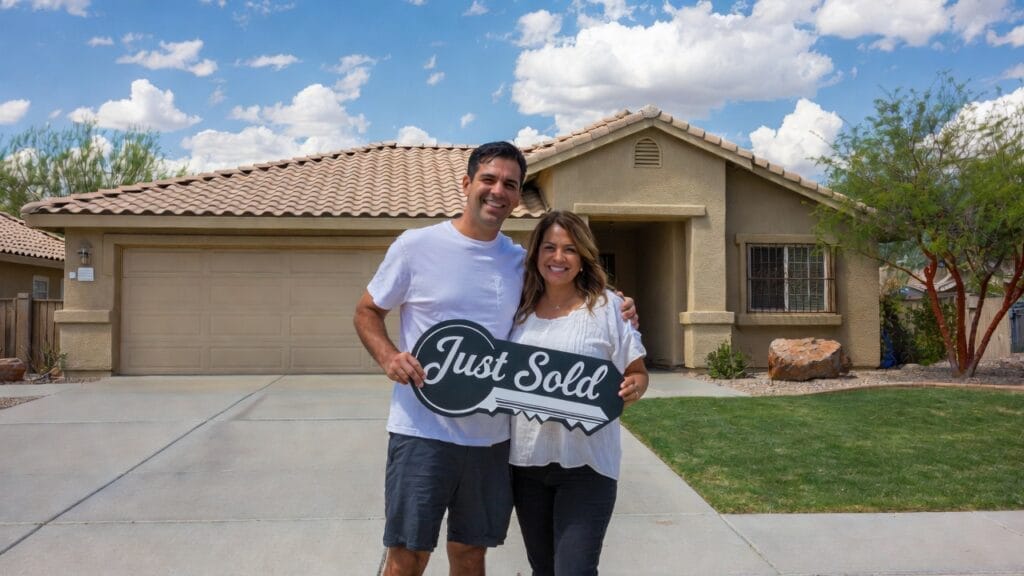 Happy couple or family standing in front of sold home with SOLD sign, Phoenix Arizona desert landscaping, mountains in background, celebrating successful home sale, warm sunset lighting, joyful expression